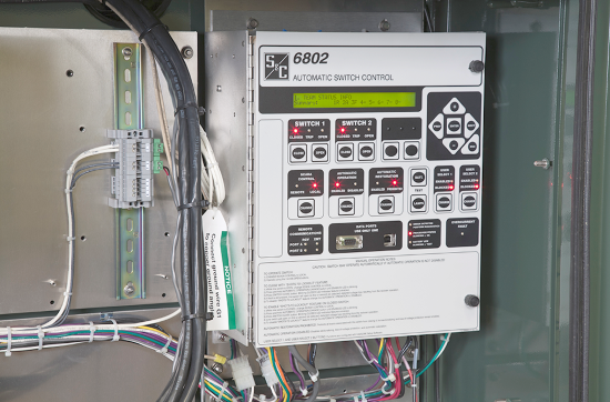 Uma unidade de Controle de Chaves 6802 montada na parede. A unidade está no lado direito da foto, enquanto o lado esquerdo mostra fios presos à parede que leva à unidade. Os fios também se projetam da parte inferior da unidade e para fora da moldura da imagem. A unidade de Controle Controle Automático de Interruptores 6802 tem um logotipo da S&C no canto superior esquerdo do painel frontal. O painel frontal tem também vários botões de touchpad rotulados; O "Botão 1" e o "Botão 2" são legíveis, todos os outros textos são muito pequenos para ler. Em alguns botões, luzes vermelhas estão acesas. 