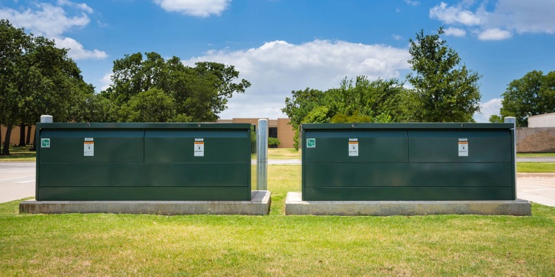 Two green underground distribution switchgear boxes on concrete pads. In the foreground is green grass. In the background are trees and a blue sky.